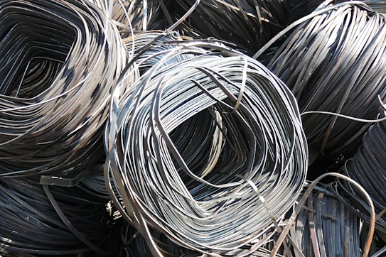 Large coils of metallic wire are tightly wound and layered together. The smooth, shiny surface of the wires reflects light prominently, creating a complex texture of curved lines. The wire is neatly stacked, giving the impression of organized industrial material ready for use.