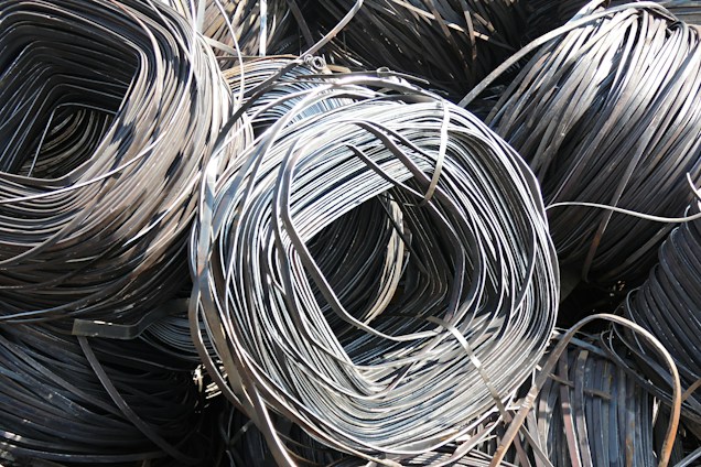 Large coils of metallic wire are tightly wound and layered together. The smooth, shiny surface of the wires reflects light prominently, creating a complex texture of curved lines. The wire is neatly stacked, giving the impression of organized industrial material ready for use.