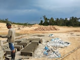 A person wearing a long-sleeved shirt, shorts, boots, and a camouflage hat is working in a construction area with concrete blocks neatly aligned on the ground. The surroundings include piles of sand or dirt, scattered tarps, and a forested area in the background under an overcast sky.