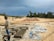 A person wearing a long-sleeved shirt, shorts, boots, and a camouflage hat is working in a construction area with concrete blocks neatly aligned on the ground. The surroundings include piles of sand or dirt, scattered tarps, and a forested area in the background under an overcast sky.