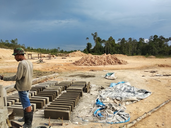 A person wearing a long-sleeved shirt, shorts, boots, and a camouflage hat is working in a construction area with concrete blocks neatly aligned on the ground. The surroundings include piles of sand or dirt, scattered tarps, and a forested area in the background under an overcast sky.