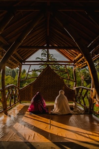 A wooden, open-air structure with a slanted roof provides a serene setting in a lush, green environment. Inside, two people sit on the wooden floor, draped in colorful cloths. The warm sunlight casts long shadows across the floor, creating a tranquil and peaceful atmosphere.