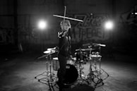 A person stands in an industrial space with graffiti on the walls, holding a drumstick in an elevated position. A drum set is positioned in the foreground with intense lights creating a dramatic ambience.