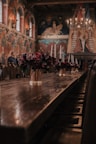 A grand, ornately decorated room with wooden tables lined with floral arrangements in vases and tall candleholders. The walls are adorned with intricate murals and tapestries, and a large chandelier with lit candles hangs from the ceiling, adding a warm glow to the space. The arched windows and medieval-style architecture evoke a historic and opulent atmosphere.