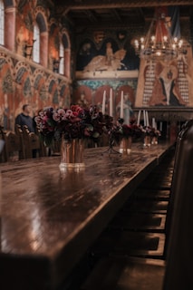 A grand, ornately decorated room with wooden tables lined with floral arrangements in vases and tall candleholders. The walls are adorned with intricate murals and tapestries, and a large chandelier with lit candles hangs from the ceiling, adding a warm glow to the space. The arched windows and medieval-style architecture evoke a historic and opulent atmosphere.