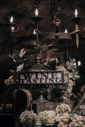 A dimly lit setting featuring a vintage chandelier with glowing candles hanging above a rustic table. Beneath it, a sign reads 'Wine Tasting' surrounded by lush floral arrangements, leaves, and decorative wine barrels. The ambiance suggests a cozy and elegant environment for a wine event.