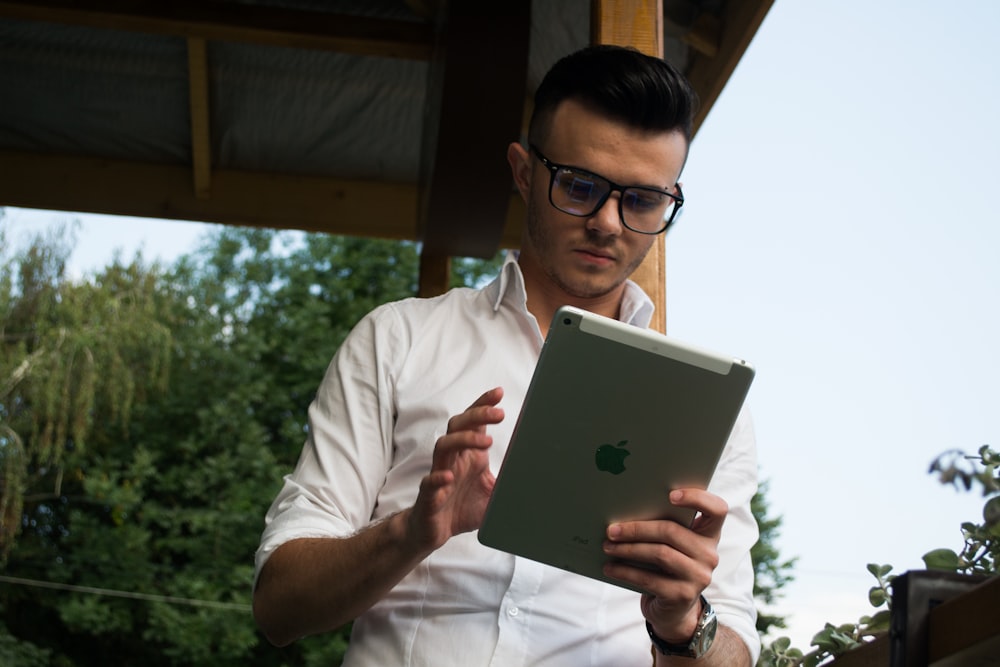 man wearing white collared button-up long-sleeved shirt and eyeglasses using iPad while standing near brown wooden post during daytime