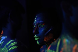 A person with neon paint on their face and body is shown in a dimly lit scene, highlighting vivid colors and patterns. The fluorescent hues contrast sharply against the dark background, creating a striking visual effect.