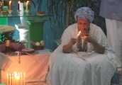 A person dressed in traditional white clothing and a headwrap is seated and lighting a candle. There is an arrangement of candles and various items like bread, flowers, and beads on a table. The setting has a serene ambiance, with green and blue hues and elements of a spiritual or ritualistic nature.