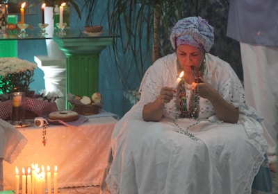 A person dressed in traditional white clothing and a headwrap is seated and lighting a candle. There is an arrangement of candles and various items like bread, flowers, and beads on a table. The setting has a serene ambiance, with green and blue hues and elements of a spiritual or ritualistic nature.