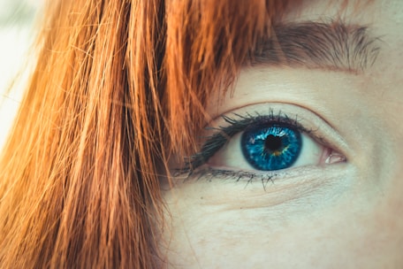 A close-up of a person's bright blue eye with intricate details of the iris visible. The surrounding area is dominated by red hair covering part of the eye and eyebrow.
