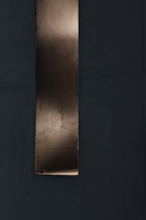 A vertical metallic strip with a reflective surface set against a dark, textured background. The metal appears slightly aged with subtle variations in tone, suggesting a weathered or industrial look.