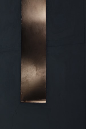 A vertical metallic strip with a reflective surface set against a dark, textured background. The metal appears slightly aged with subtle variations in tone, suggesting a weathered or industrial look.