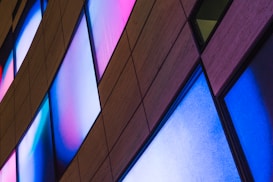 A series of diamond-shaped panels with glowing colors in shades of blue, pink, and white positioned on a building facade. The panels are arranged in a diagonal pattern against a textured, wooden background.