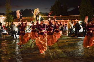 A group of people performing a traditional dance in an outdoor setting at night. The dancers are wearing burgundy and orange costumes and are holding decorative objects in their hands, possibly feathers or fans. The scene is lively with spectators gathered around, and there's a backdrop of trees and buildings.