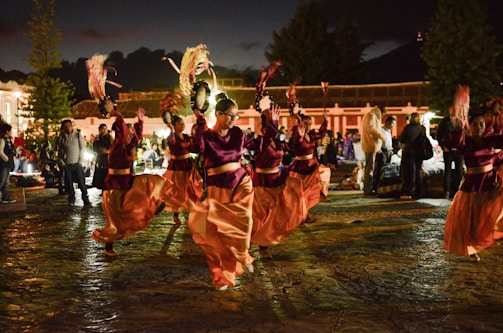 A group of people performing a traditional dance in an outdoor setting at night. The dancers are wearing burgundy and orange costumes and are holding decorative objects in their hands, possibly feathers or fans. The scene is lively with spectators gathered around, and there's a backdrop of trees and buildings.