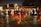 A group of people performing a traditional dance in an outdoor setting at night. The dancers are wearing burgundy and orange costumes and are holding decorative objects in their hands, possibly feathers or fans. The scene is lively with spectators gathered around, and there's a backdrop of trees and buildings.