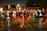 A group of people performing a traditional dance in an outdoor setting at night. The dancers are wearing burgundy and orange costumes and are holding decorative objects in their hands, possibly feathers or fans. The scene is lively with spectators gathered around, and there's a backdrop of trees and buildings.