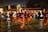 A group of people performing a traditional dance in an outdoor setting at night. The dancers are wearing burgundy and orange costumes and are holding decorative objects in their hands, possibly feathers or fans. The scene is lively with spectators gathered around, and there's a backdrop of trees and buildings.
