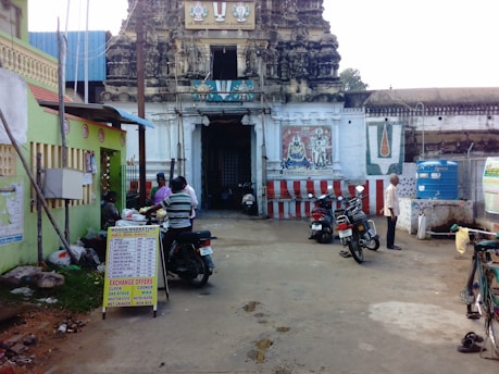 A temple entrance with intricately carved stone architecture is visible. Several motorcycles are parked nearby, and people are engaged in conversation or walking. Signage lists various market items such as kitchen appliances. A large painted mural is on the temple wall accompanied by a religious symbol. Nearby, there is a water storage tank and a bicycle.