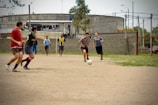 A group of men are playing soccer on a dirt field. The players are in casual clothing, with no uniforms, indicating a friendly or informal match. In the background, there is an industrial-looking building with laundry hanging outside, and other people can be seen watching the game.