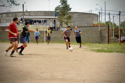 A group of men are playing soccer on a dirt field. The players are in casual clothing, with no uniforms, indicating a friendly or informal match. In the background, there is an industrial-looking building with laundry hanging outside, and other people can be seen watching the game.