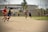 A group of men are playing soccer on a dirt field. The players are in casual clothing, with no uniforms, indicating a friendly or informal match. In the background, there is an industrial-looking building with laundry hanging outside, and other people can be seen watching the game.