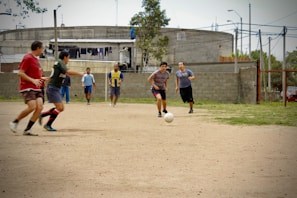 A group of men are playing soccer on a dirt field. The players are in casual clothing, with no uniforms, indicating a friendly or informal match. In the background, there is an industrial-looking building with laundry hanging outside, and other people can be seen watching the game.