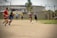 A group of men are playing soccer on a dirt field. The players are in casual clothing, with no uniforms, indicating a friendly or informal match. In the background, there is an industrial-looking building with laundry hanging outside, and other people can be seen watching the game.