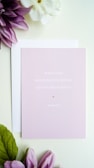 A light pink card with the text 'What God has joined together, let no one separate. Mark 10:9' is placed on top of a white envelope. Surrounding the card are purple and white flowers with green leaves, all set against a soft white background.