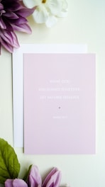 A light pink card with the text 'What God has joined together, let no one separate. Mark 10:9' is placed on top of a white envelope. Surrounding the card are purple and white flowers with green leaves, all set against a soft white background.
