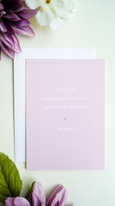 A light pink card with the text 'What God has joined together, let no one separate. Mark 10:9' is placed on top of a white envelope. Surrounding the card are purple and white flowers with green leaves, all set against a soft white background.