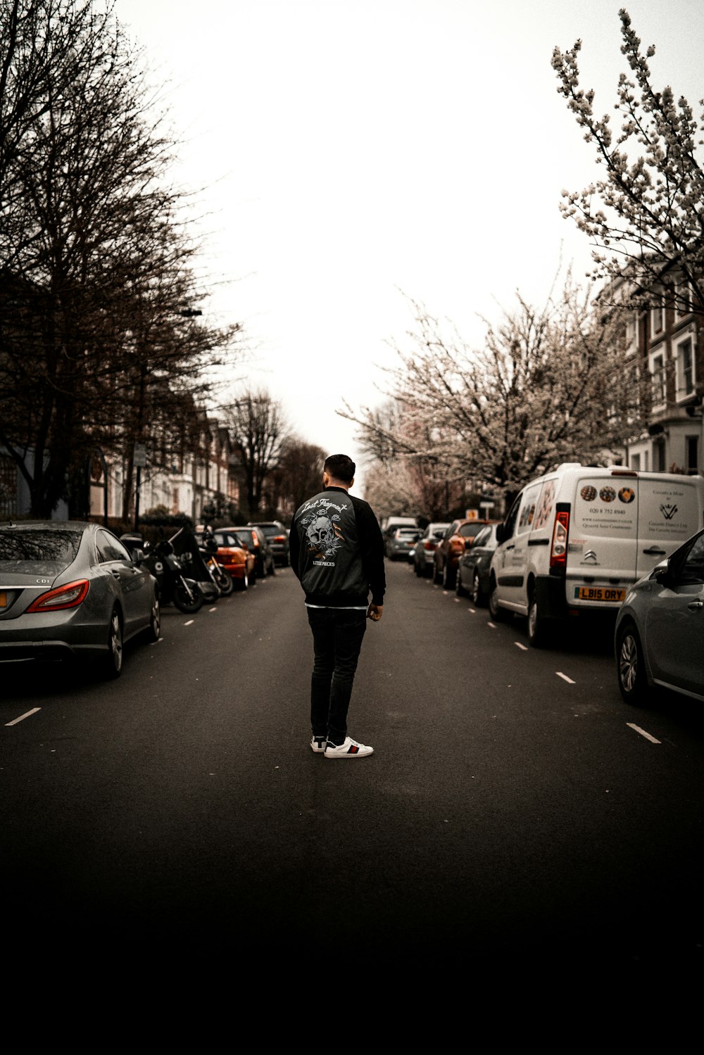 man in black jacket standing at the center of the road between vehicle parked on road side