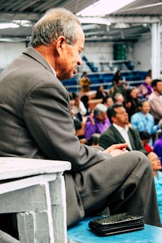 An elderly man in a suit is seated on bleachers, surrounded by a crowd of people. The setting appears to be indoors, possibly a community event or gathering. His facial expression is contemplative, and he holds something in his hands. Other attendees, of various ages and genders, are seated in the background.