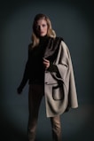 A person stands confidently wearing a fashionable ensemble. The outfit includes a dark turtleneck sweater and a stylish gray coat with a fur accent draped over one shoulder. The background is a muted, dark color, contributing to an elegant and sophisticated aura.