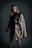 A person stands confidently wearing a fashionable ensemble. The outfit includes a dark turtleneck sweater and a stylish gray coat with a fur accent draped over one shoulder. The background is a muted, dark color, contributing to an elegant and sophisticated aura.