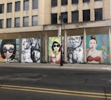 A series of colorful street art murals are displayed on the side of an urban building. The artwork features large, expressive portraits of people, some holding lollipops. The backgrounds of the murals include vibrant geometric patterns. A vertical black streetlight is situated in front of the murals.