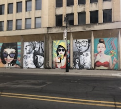 A series of colorful street art murals are displayed on the side of an urban building. The artwork features large, expressive portraits of people, some holding lollipops. The backgrounds of the murals include vibrant geometric patterns. A vertical black streetlight is situated in front of the murals.