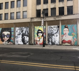 A series of colorful street art murals are displayed on the side of an urban building. The artwork features large, expressive portraits of people, some holding lollipops. The backgrounds of the murals include vibrant geometric patterns. A vertical black streetlight is situated in front of the murals.