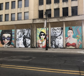 A series of colorful street art murals are displayed on the side of an urban building. The artwork features large, expressive portraits of people, some holding lollipops. The backgrounds of the murals include vibrant geometric patterns. A vertical black streetlight is situated in front of the murals.