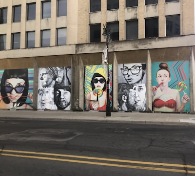 A series of colorful street art murals are displayed on the side of an urban building. The artwork features large, expressive portraits of people, some holding lollipops. The backgrounds of the murals include vibrant geometric patterns. A vertical black streetlight is situated in front of the murals.