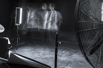 A dimly lit studio scene featuring blurred motion of a person against a dark background. The setup includes professional lighting equipment and a large industrial fan. The overall ambiance is experimental and surreal, with faint, ghostly figures adding a sense of movement and mystery.