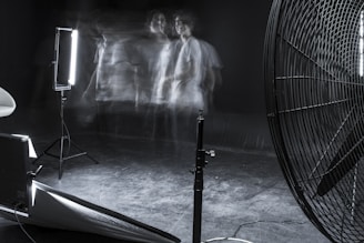 A dimly lit studio scene featuring blurred motion of a person against a dark background. The setup includes professional lighting equipment and a large industrial fan. The overall ambiance is experimental and surreal, with faint, ghostly figures adding a sense of movement and mystery.