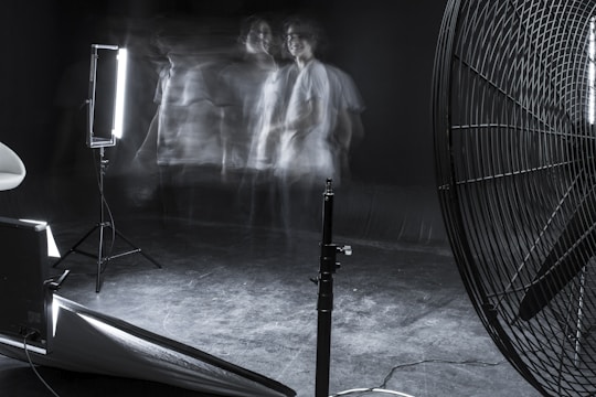 A dimly lit studio scene featuring blurred motion of a person against a dark background. The setup includes professional lighting equipment and a large industrial fan. The overall ambiance is experimental and surreal, with faint, ghostly figures adding a sense of movement and mystery.