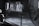 A dimly lit studio scene featuring blurred motion of a person against a dark background. The setup includes professional lighting equipment and a large industrial fan. The overall ambiance is experimental and surreal, with faint, ghostly figures adding a sense of movement and mystery.