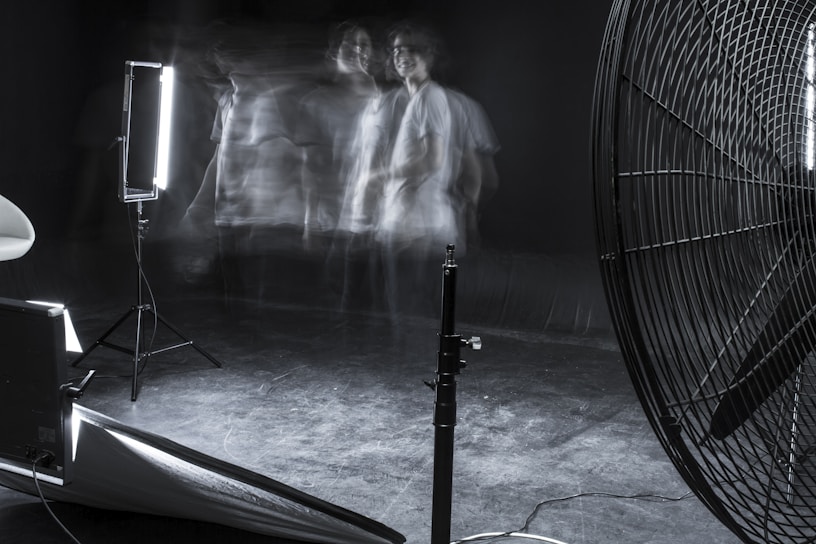A dimly lit studio scene featuring blurred motion of a person against a dark background. The setup includes professional lighting equipment and a large industrial fan. The overall ambiance is experimental and surreal, with faint, ghostly figures adding a sense of movement and mystery.
