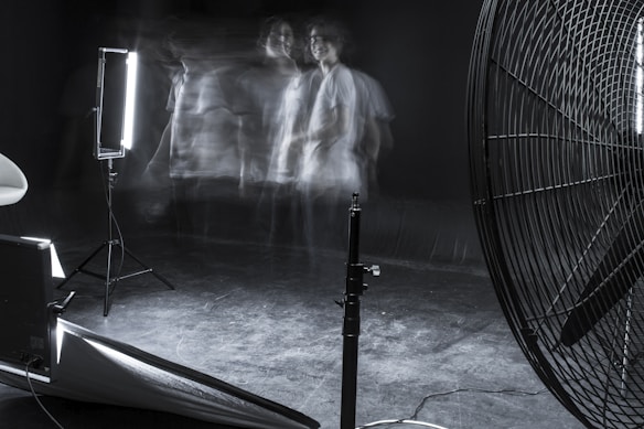 A dimly lit studio scene featuring blurred motion of a person against a dark background. The setup includes professional lighting equipment and a large industrial fan. The overall ambiance is experimental and surreal, with faint, ghostly figures adding a sense of movement and mystery.
