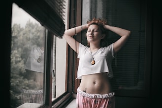 A relaxed individual stands in a contemplative pose by a window, wearing casual clothes with a pendant necklace. The person has an expression of serenity and is framed by the natural light coming from outside, where greenery is visible.