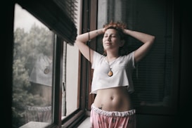 A relaxed individual stands in a contemplative pose by a window, wearing casual clothes with a pendant necklace. The person has an expression of serenity and is framed by the natural light coming from outside, where greenery is visible.