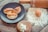 A dark plate holds three baked pastries filled with a white creamy substance. Nearby, a rolling pin is placed on a wooden surface dusted with flour, next to a whole brown egg. A wooden round container with a dollop of cream and some seeds is also visible.